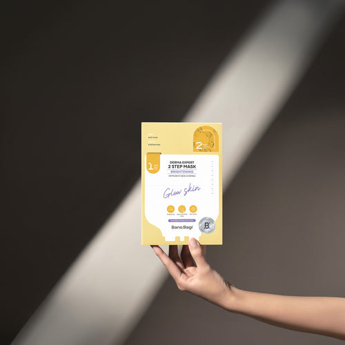 Hand holding a Bano Bagi Derma Expert 2 Step Mask Brightening against a dark background with a ray of sunshine
