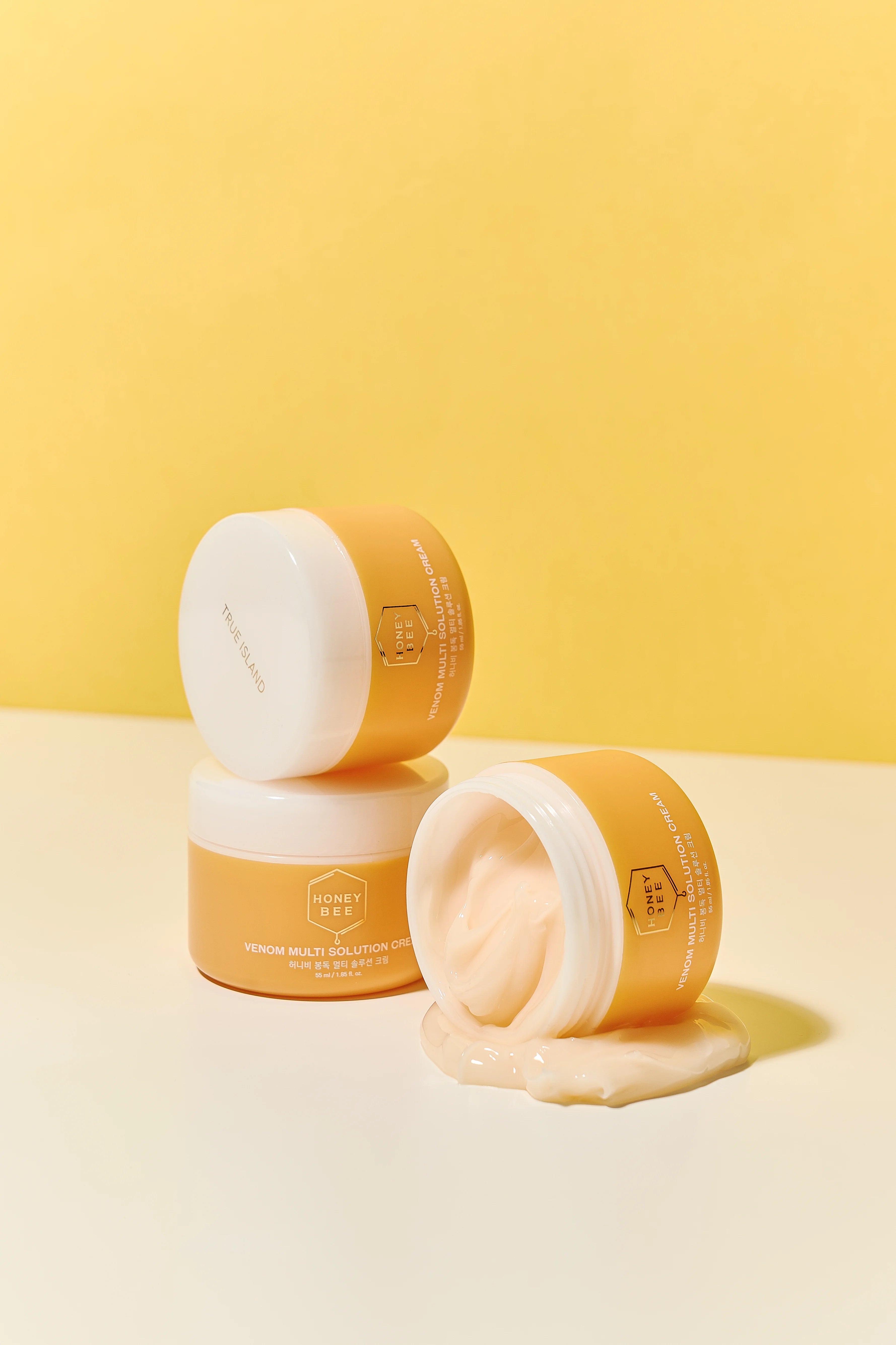 Three Honey Bee Venom Multi Solution Creams on a white surface against a yellow background, with one jar tipped over so the product pools on the tabletop