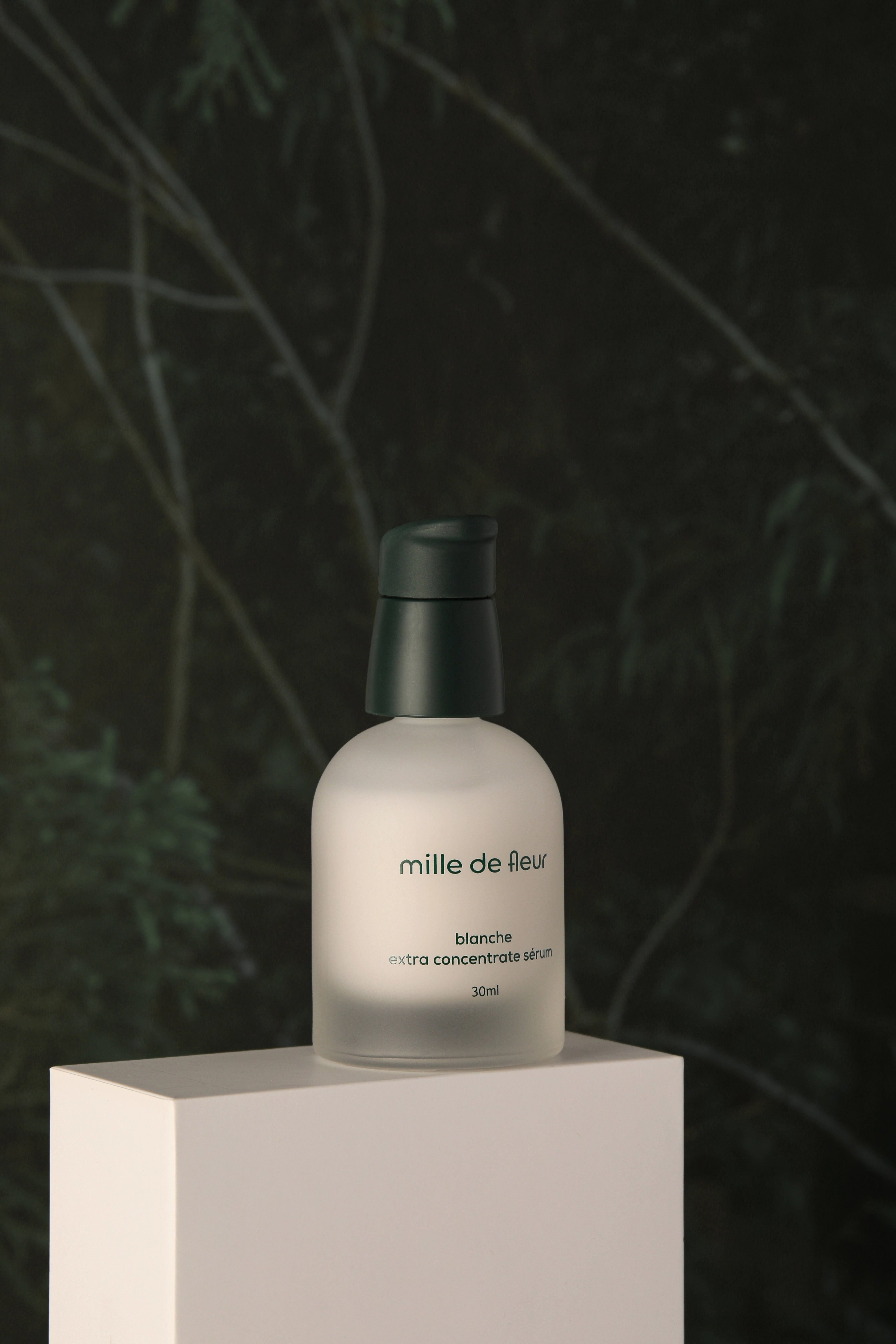 Mille de Fleur Blanche Extra Concentrate Sérum on a white platform with a dark green leafy background