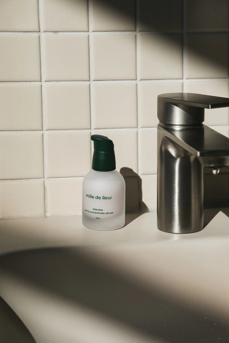 Bottle of Mille de Fleur Blanche Extra Concentrate Sérum on a bathroom counter with a tiled wall in the background